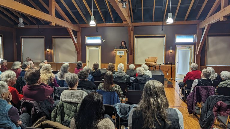 A woman stands at a podium speaking to a seated audience in a wooden hall with exposed beams, large windows, and overhead lights.