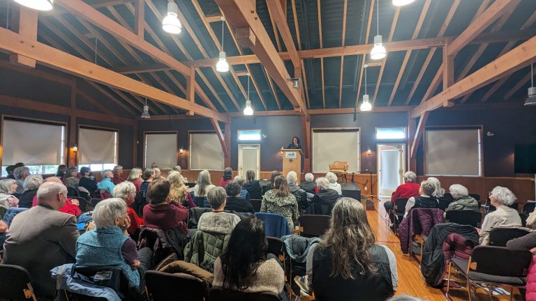 A speaker stands at a podium addressing an audience seated in a wooden hall with exposed beams and large windows.