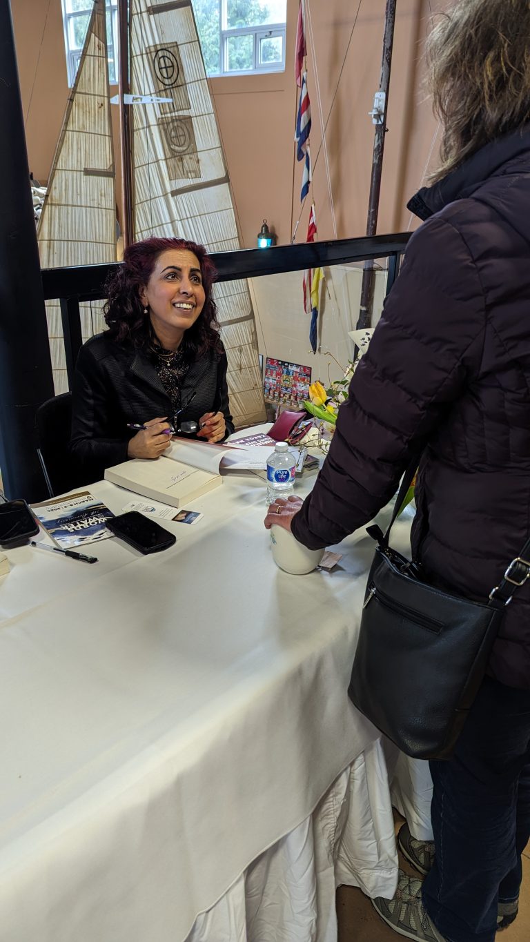 A smiling woman sits at a table signing a book for another person, who is standing and wearing a dark jacket. Flowers, books, and brochures are on the table. Sail-themed decor and flags are visible in the background.