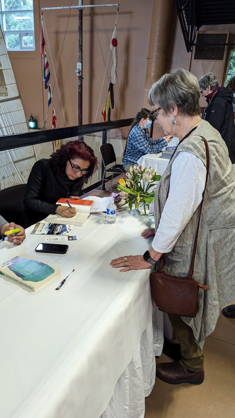 An author signs a book for an older woman at a table covered with white cloth, with other people and books in the background.