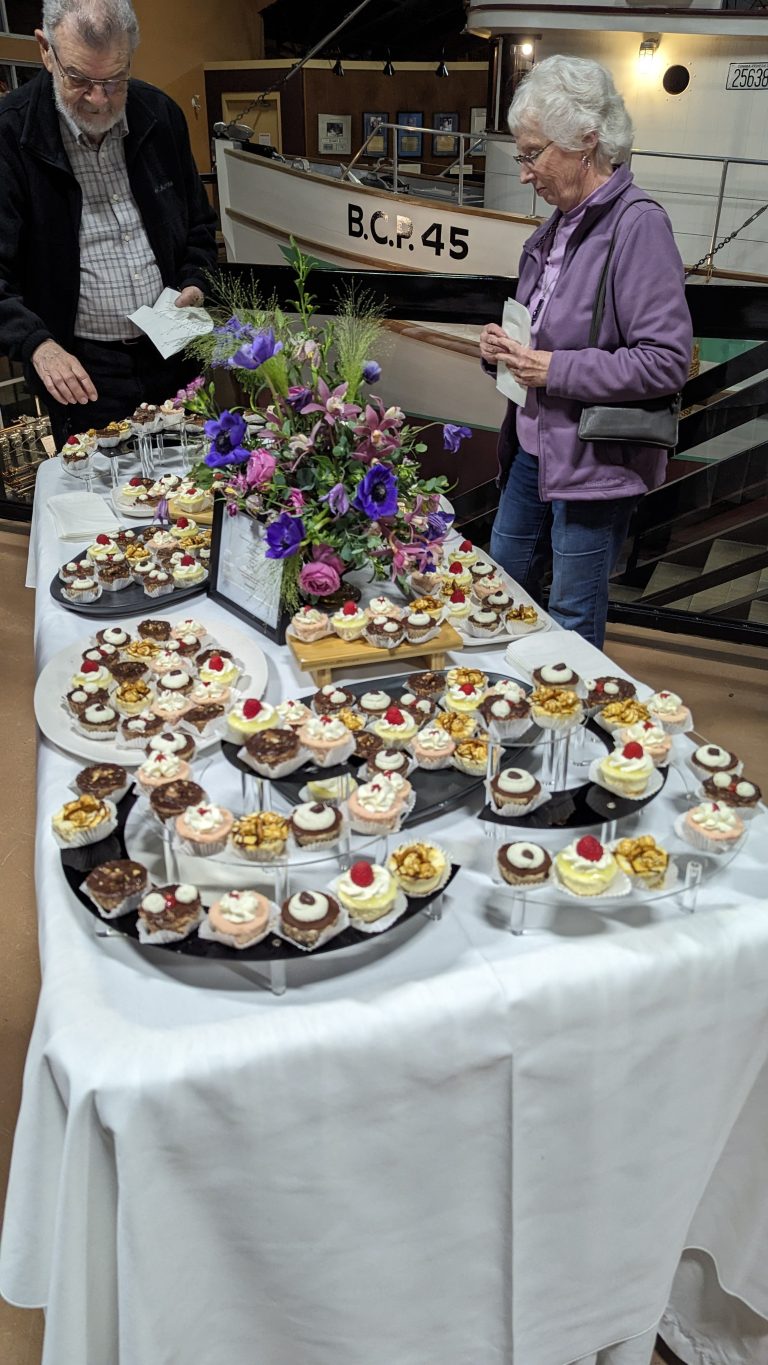 A table covered with a white tablecloth displays assorted mini desserts and a flower arrangement. Two older adults stand nearby, selecting treats. A boat is visible in the background.