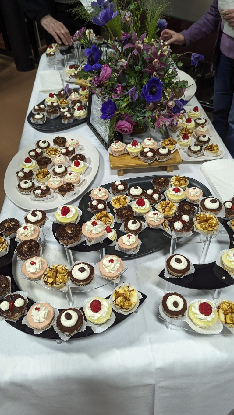 A table covered with assorted mini desserts, including cupcakes with cream, nuts, and raspberries, arranged around a floral centerpiece. Several hands are reaching for treats.