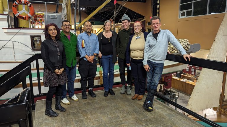 Seven people stand together indoors on a balcony, smiling at the camera. Behind them are nautical displays, ropes, and a large lifesaver labeled “B.C. Coast Vance.”.