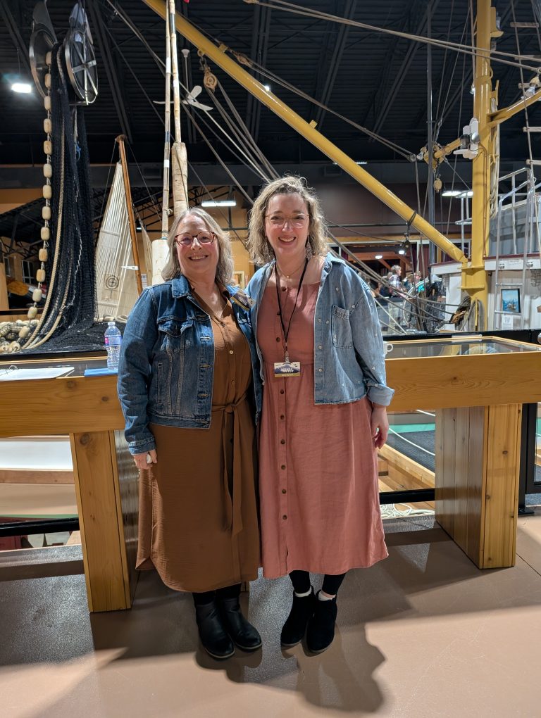 Two women stand smiling indoors by a wooden railing, both wearing brown dresses and denim jackets, with boats and nautical equipment displayed in the background.