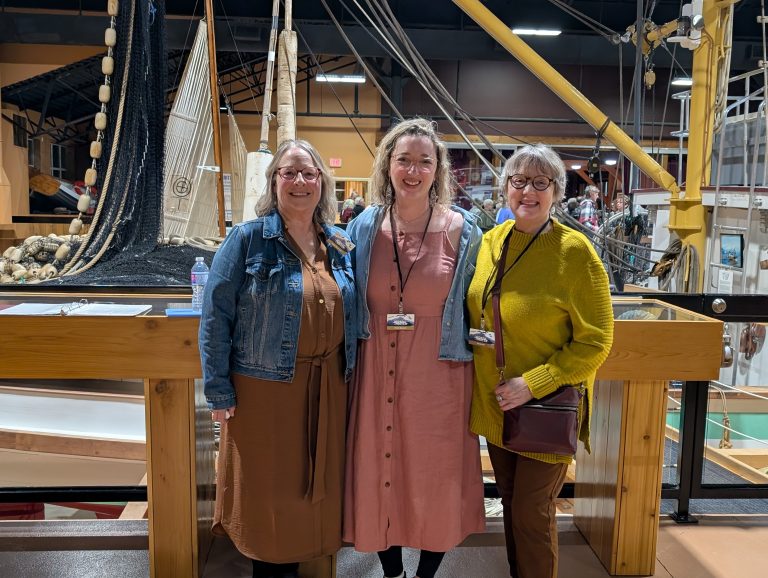 Three women stand smiling in front of a wooden railing inside a maritime museum, with ship models, fishing nets, and nautical equipment displayed in the background.