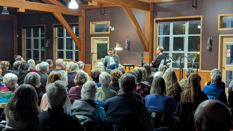 A large audience listens to two people having a discussion onstage in a wood-paneled room with large windows and exposed beams. The setting appears to be a public talk or interview.