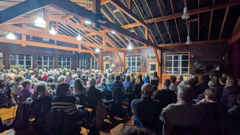 A large audience sits in rows inside a wooden hall, attentively watching a person speak or perform at the front stage under bright ceiling lights.