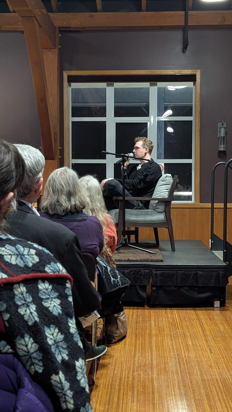 A person sits on a chair on a small stage, speaking into a microphone. Audience members face the stage, listening attentively in a wood-paneled room with large windows behind the speaker.