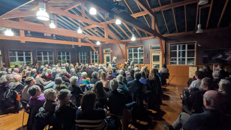 A large group of people sit in rows of chairs inside a wooden hall, attentively watching a person speaking at the front under bright ceiling lights.