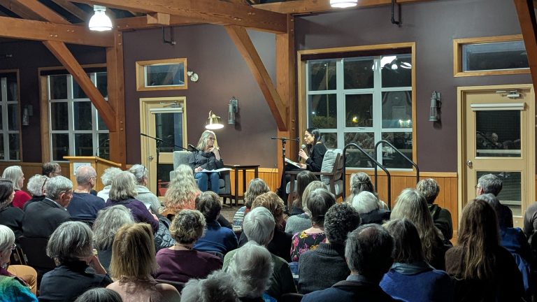Two women sit on a stage having a conversation in front of a seated audience in a wooden-beamed room with large windows. Microphones and lamps are set up for the discussion.