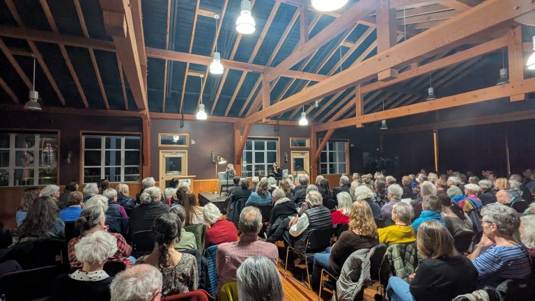 A large audience sits in a wooden hall with exposed beams, watching a person speak at a podium under bright lights. The room is filled with people of varying ages, all facing the stage.