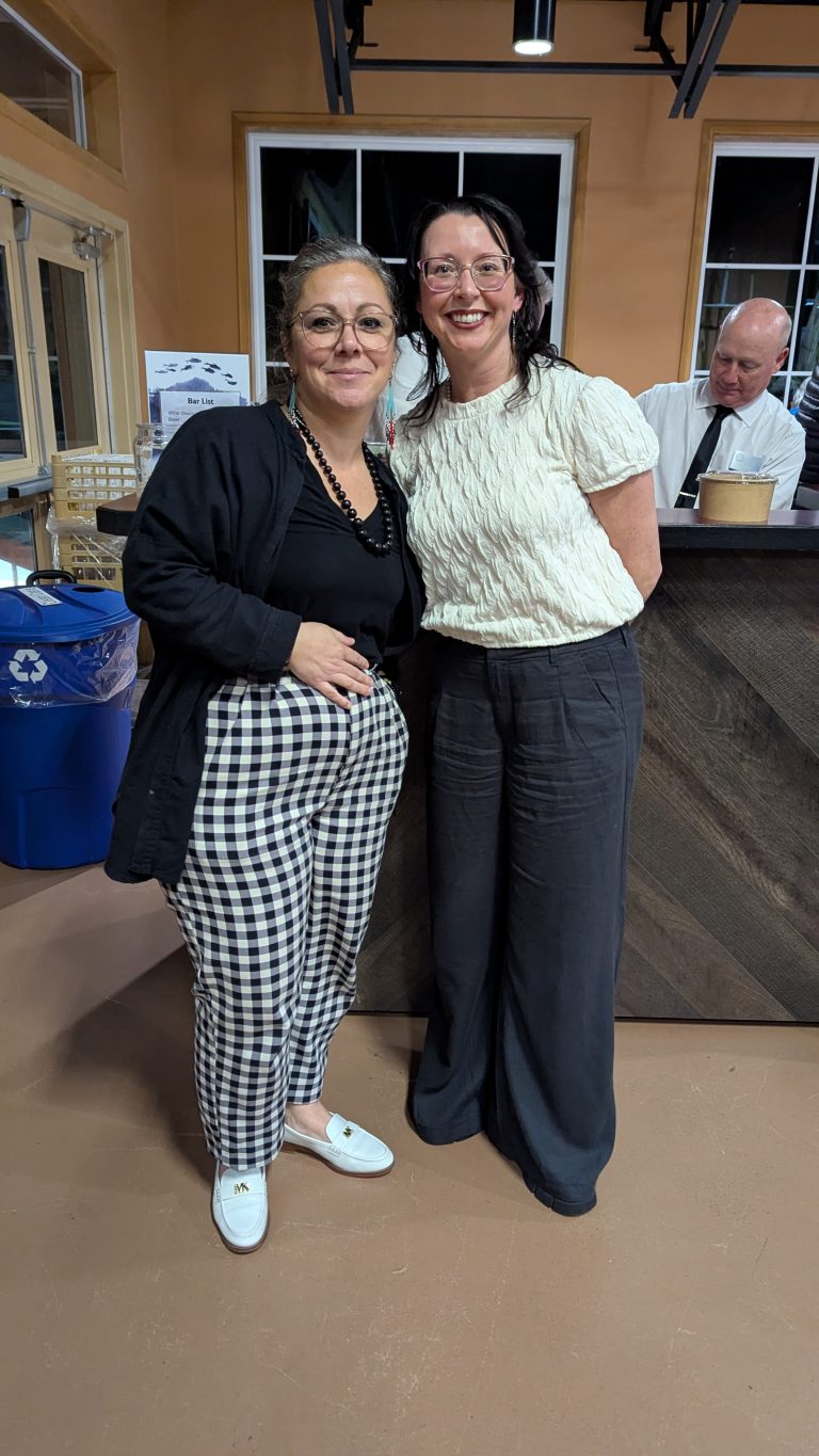 Two women stand side by side smiling indoors near a counter, with a man in a white shirt behind them. One wears checkered pants and glasses; the other wears a textured white top with dark pants.