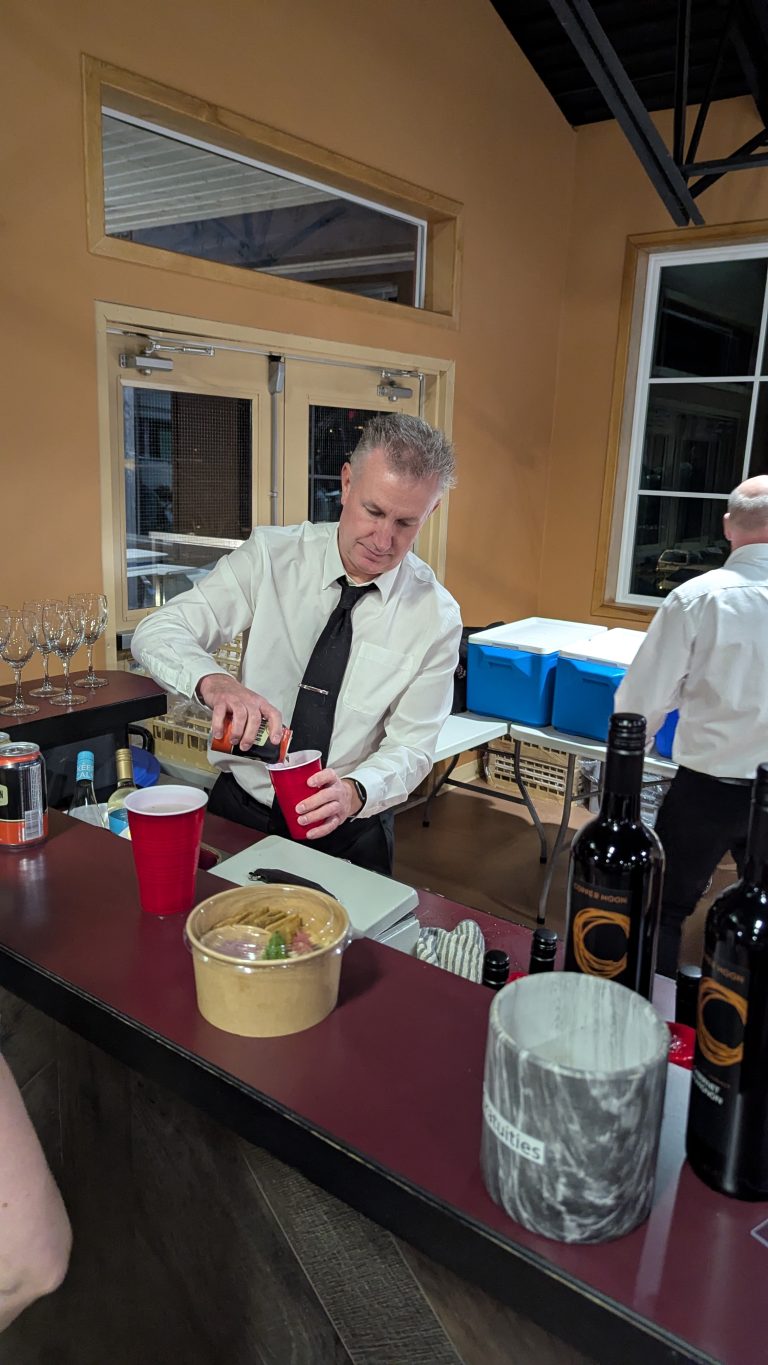 A bartender in a white shirt and black tie pours a drink into a red cup at a bar, with wine bottles, glasses, and food nearby.