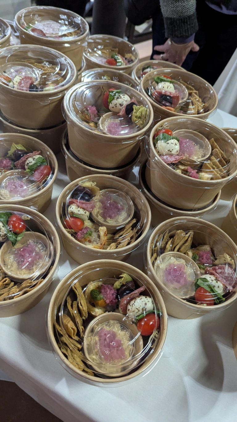 Stacks of round brown paper bowls with clear lids, filled with colorful appetizers including cherry tomatoes, crackers, cheese, and pickled onions, arranged on a white tablecloth.