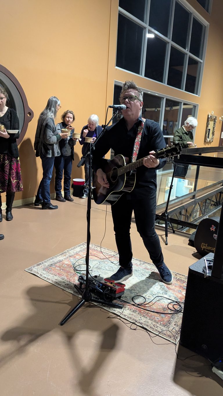A musician in black sings and plays guitar into a microphone on a small indoor stage. Several people in the background watch, chat, and hold drinks. The setting appears to be a casual indoor gathering.