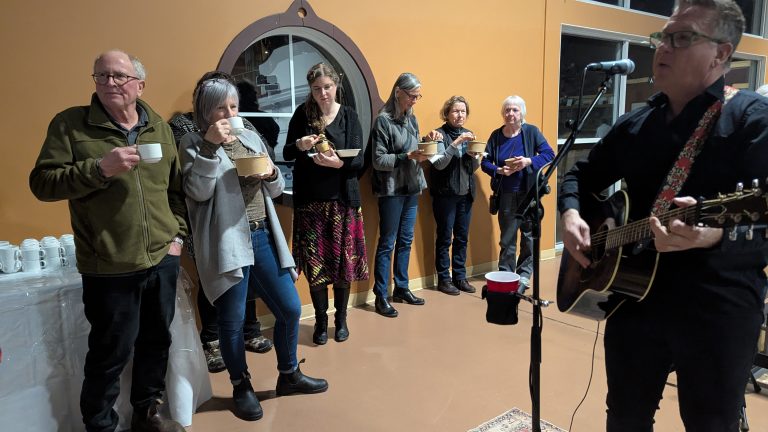 A group of people stand indoors, eating and drinking, while a man in glasses plays guitar and sings at a microphone. Cups and food containers are visible; the mood appears relaxed and social.