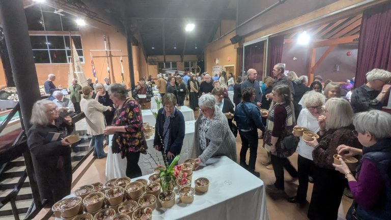 A large group of people gathers indoors, serving themselves food from bowls on tables, engaging in conversation in a warmly lit event space.