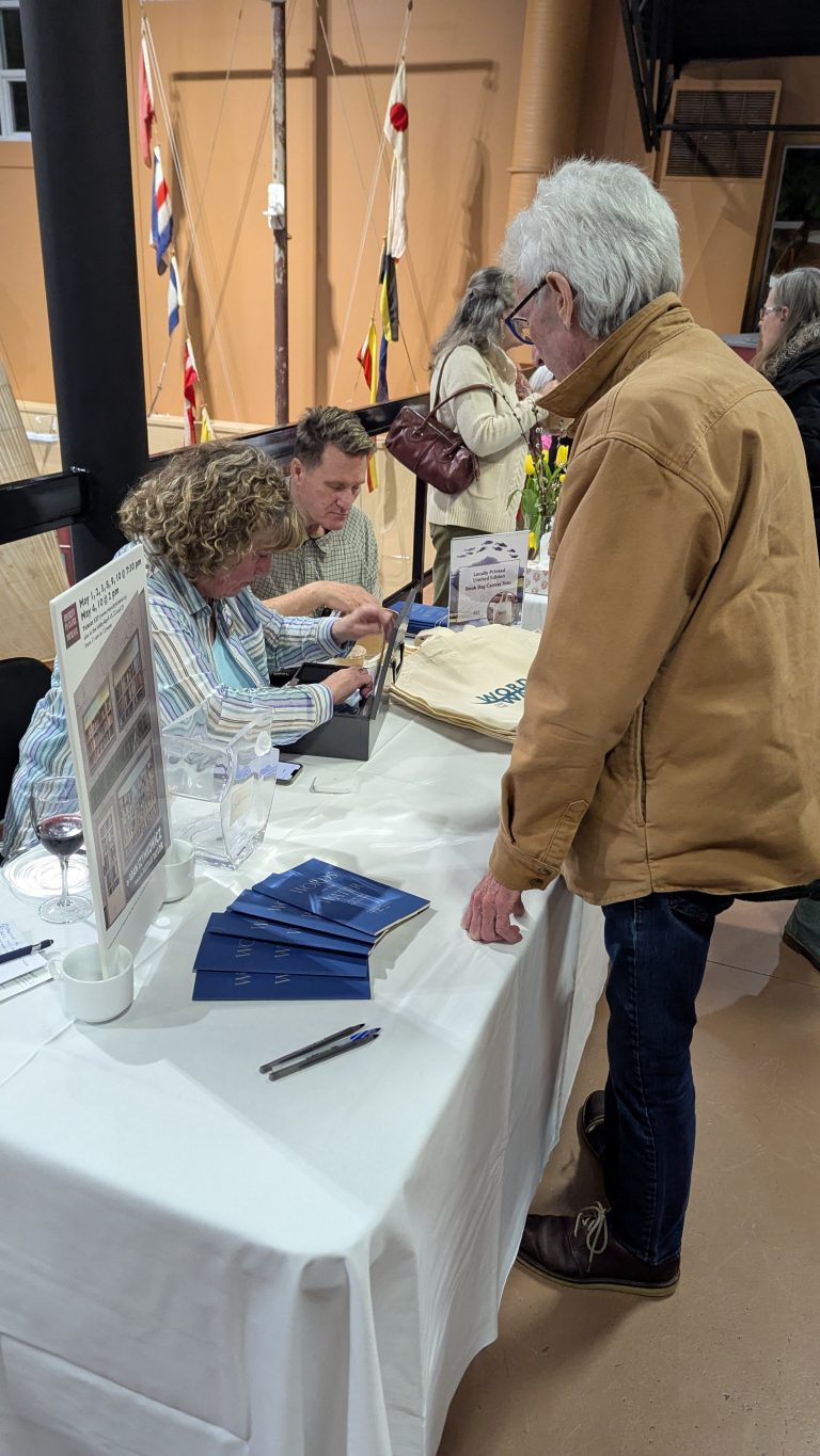 Two people sit at a table signing books while a man in a tan jacket stands in front of them. Pamphlets, a glass, and a tote bag are on the table. Other people are in the background near nautical flags.