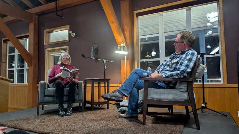 Two people sit and talk on a stage in a cozy room; one reads from a book while the other listens. Large windows, wooden beams, and lamps are visible in the background.