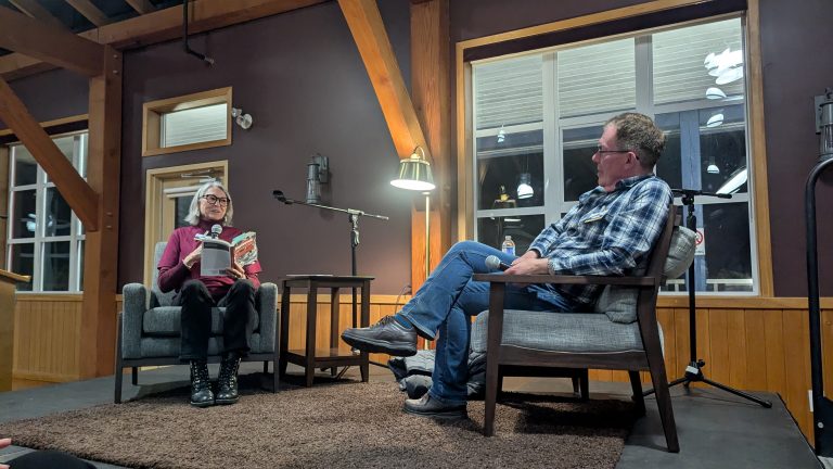 Two people sit facing each other on a stage in a cozy room; one holds an open book and reads aloud, while the other listens attentively. Microphones and small tables separate them.