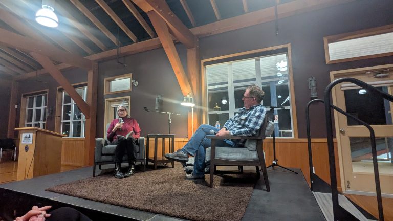 Two people sit on a stage in armchairs, having a conversation. The room has exposed wooden beams, large windows, and warm lighting. One person holds a microphone while the other listens.