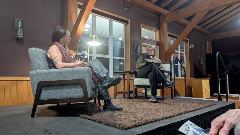 Two people sit facing each other on stage in armchairs, having a conversation. One holds a microphone, the other holds a notepad. The setting appears to be a cozy, wood-paneled room with large windows.