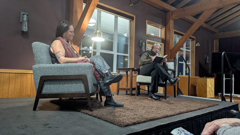 Two people sit on stage in armchairs, engaged in conversation. One reads from a book. The setting is indoors, with wooden beams and large windows in the background.