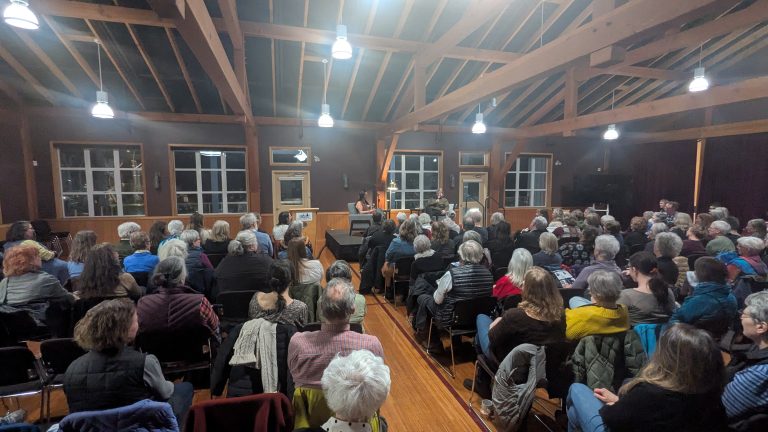 A large audience sits in a wooden hall, facing a stage where two people are having a discussion. The room is well-lit with exposed beams and large windows.