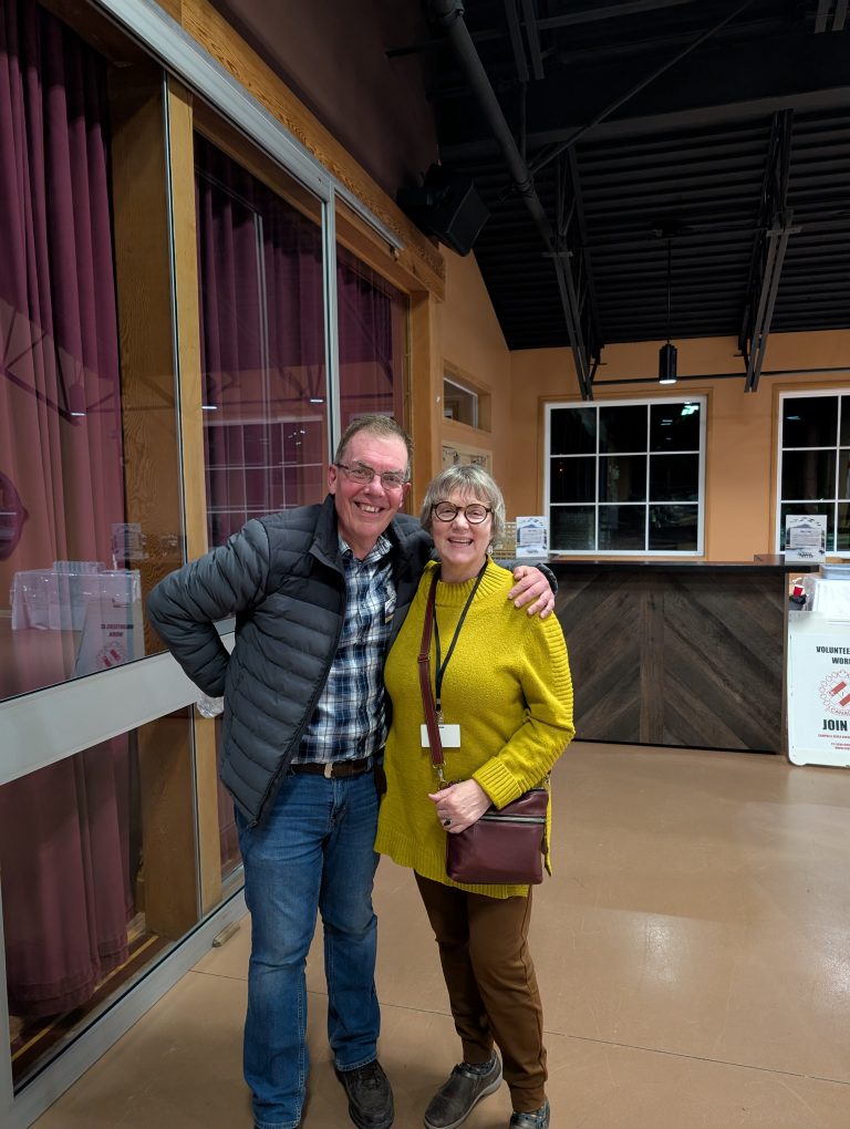 Two people smile and pose together indoors by a glass door and a counter, with maroon curtains in the background. One wears a gray jacket; the other wears a yellow sweater and glasses.