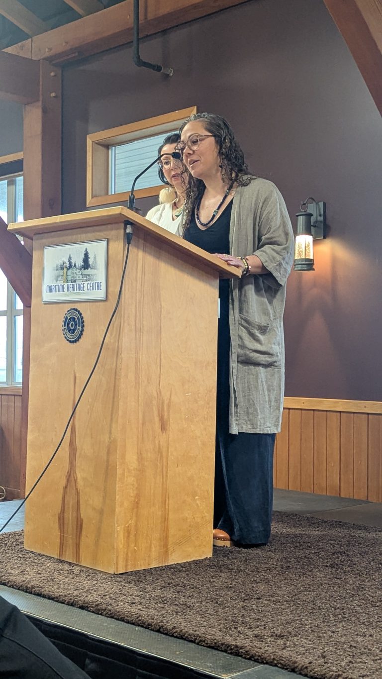 Two women stand at a wooden podium labeled "Maritime Heritage Centre," speaking into a microphone in a room with wood paneling and beams.
