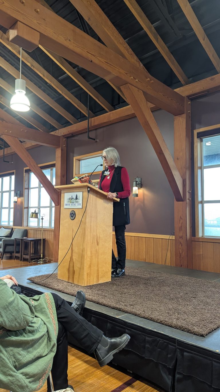 A woman with gray hair, wearing a red top and black vest, speaks at a wooden podium on a small stage in a rustic room with exposed beams and large windows. People are seated and listening.