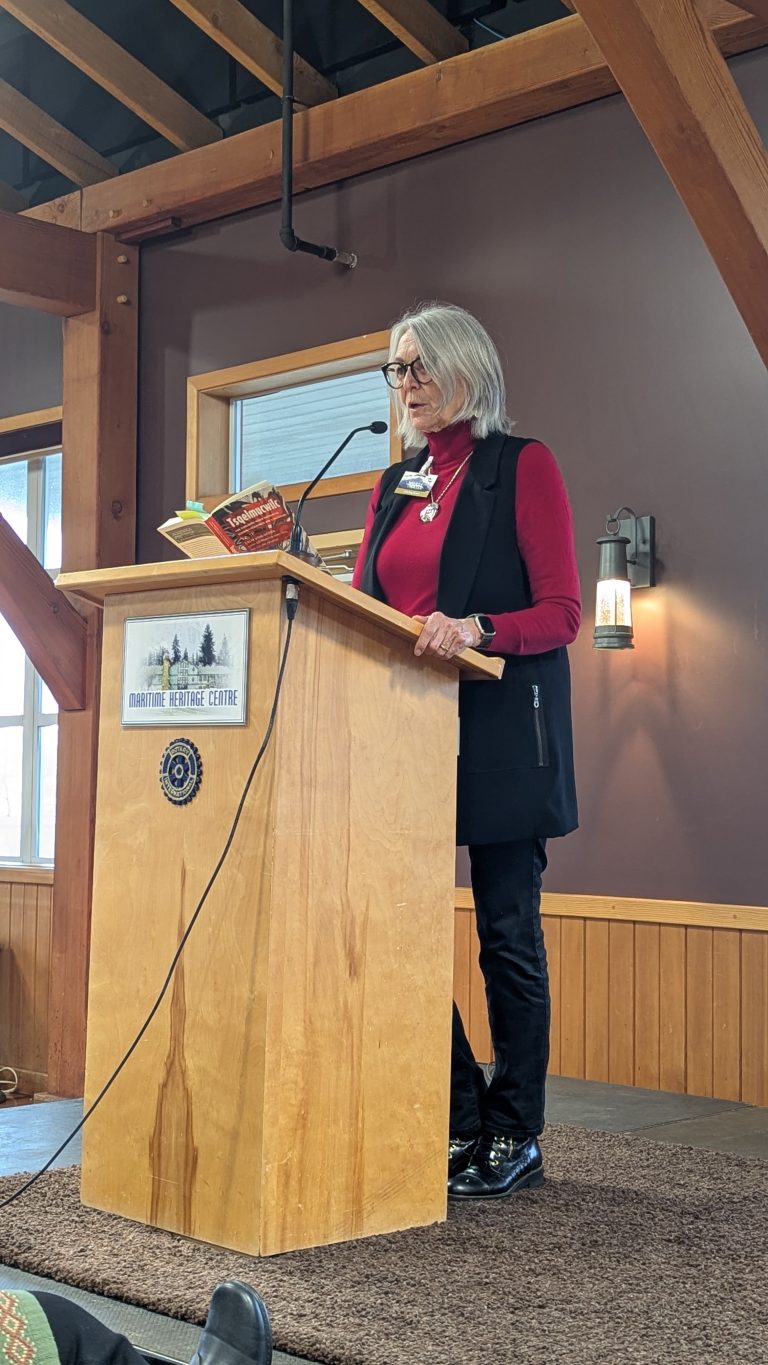 A person with gray hair, glasses, and a red long-sleeve top speaks at a wooden podium inside a building with wood beams. They are holding a book and wearing a black vest and pants.