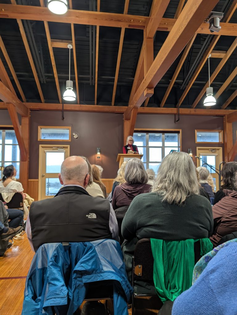 A woman speaks at a podium in a wooden-beamed room, facing an audience seated on chairs. People listen attentively, some with gray hair, wearing jackets and sweaters. Large windows are in the background.