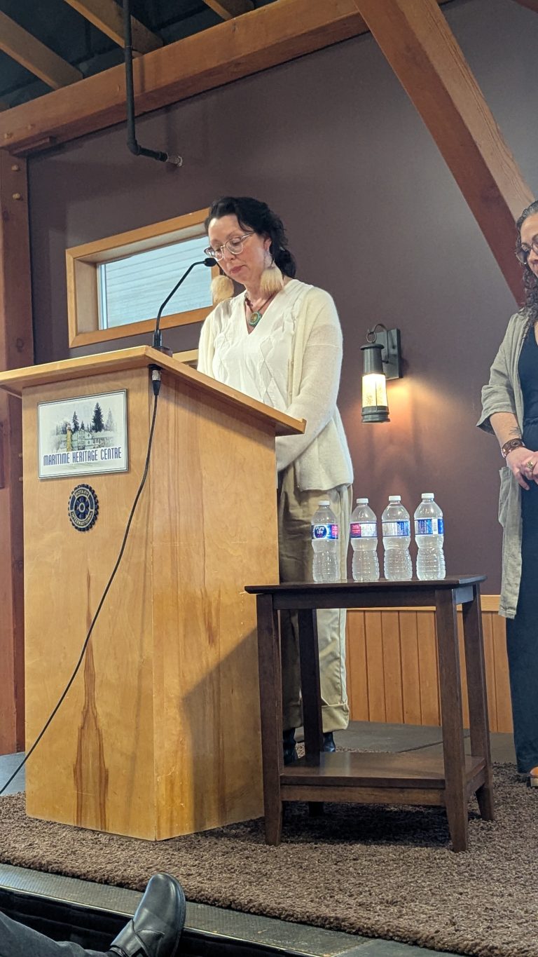 A woman stands at a wooden podium, reading from papers. Four water bottles are on a small table beside her. Another person stands nearby. The setting appears to be indoors, at the Maritime Heritage Centre.
