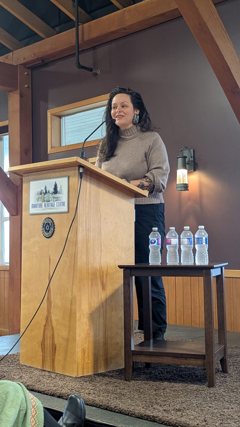 A woman with long dark hair speaks at a wooden podium indoors. Four water bottles sit on a small table beside her. The room has wooden beams, a window, and a wall lamp.