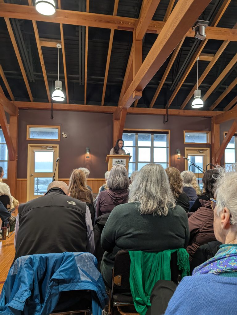 A person stands at a podium speaking to a seated audience in a wood-beamed room with large windows. The crowd listens attentively, facing the speaker at the front of the room.