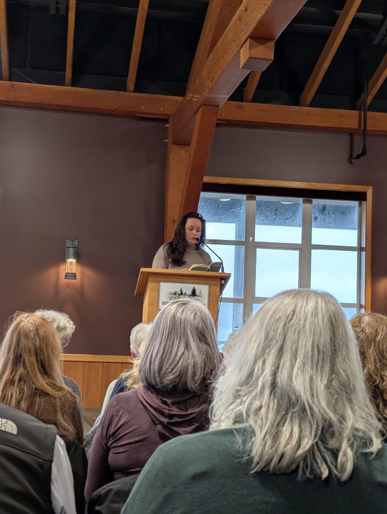 A person stands at a podium reading from a book to an audience in a room with wooden beams and a large window. The audience is facing forward, listening attentively.