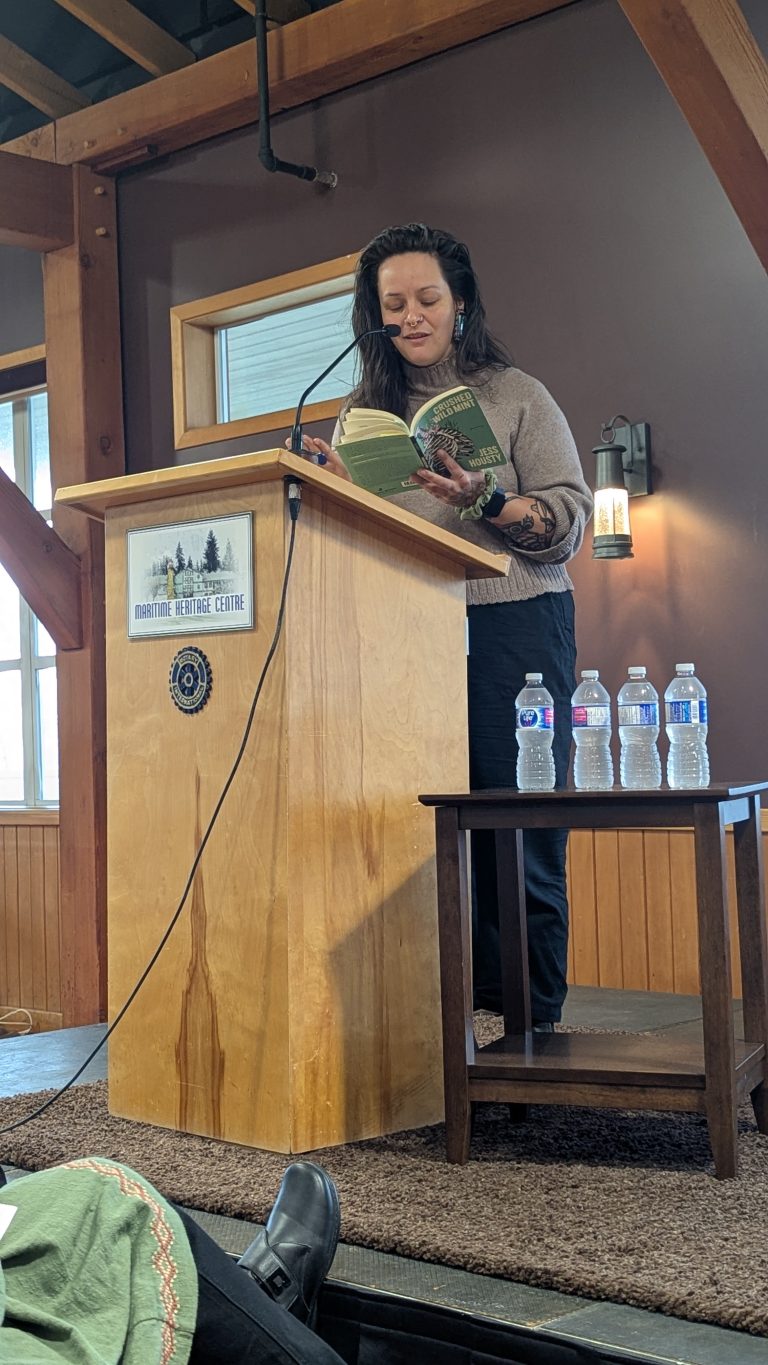 A woman stands at a wooden podium reading from a book in a room with wood beams. Three water bottles sit on a small table nearby. A window lets in natural light behind her.