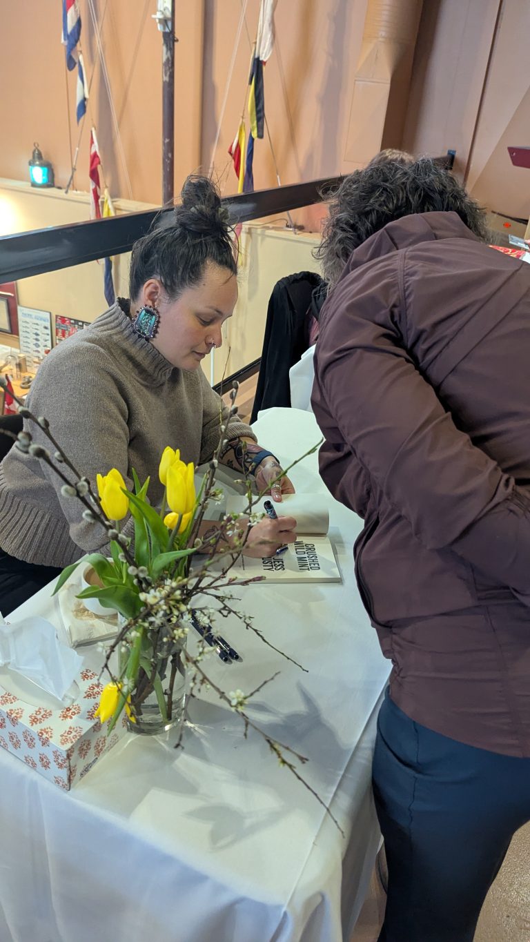 A woman sits at a table with yellow tulips, signing a book for another person standing across from her at an indoor event.