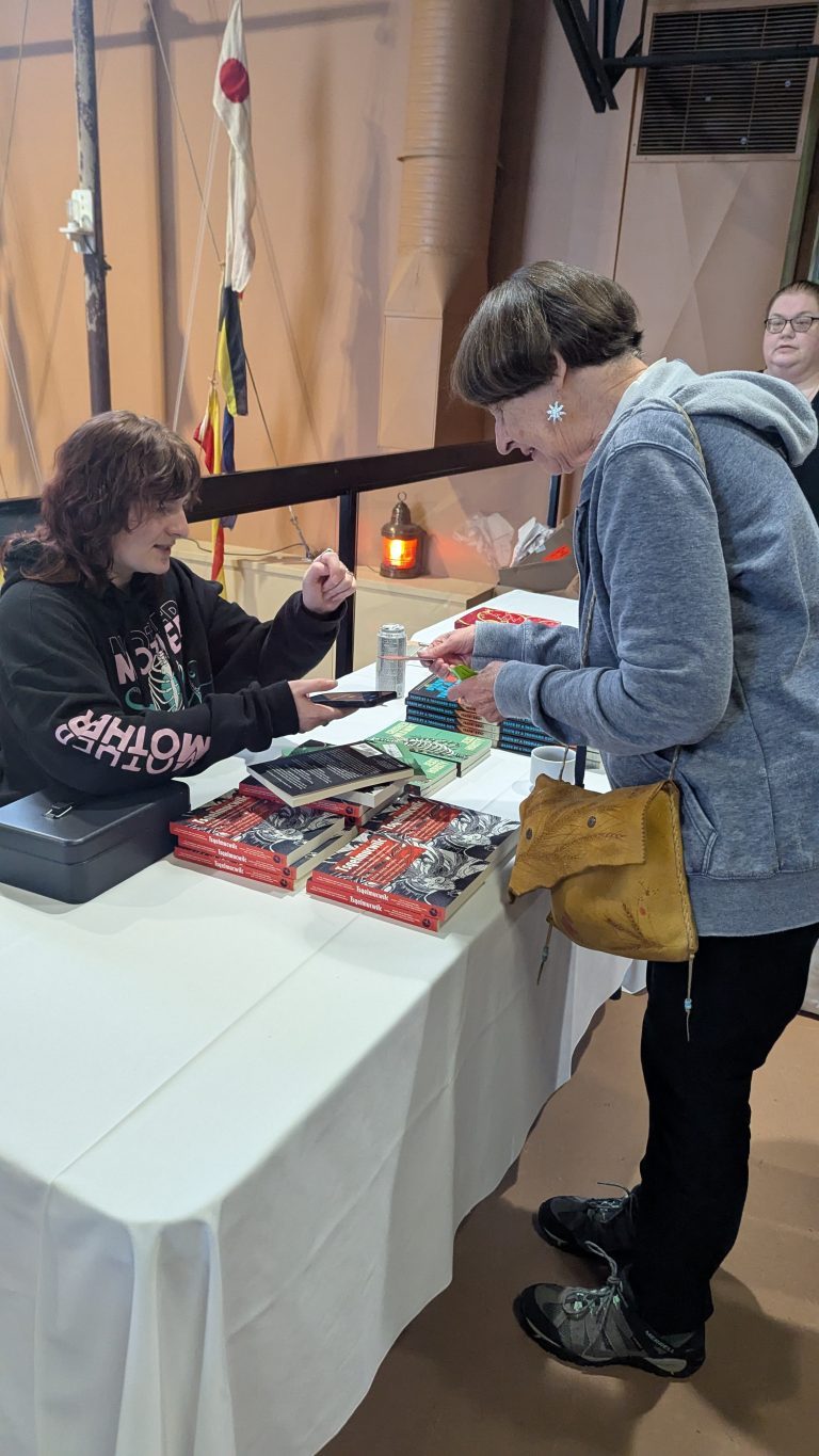 A woman in a gray hoodie buys books from a vendor at a table covered with stacks of red books and crocheted items, inside a building with nautical decor.