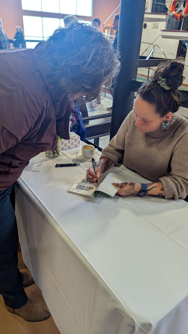 A woman sits at a table signing a book for a standing person. The table has a white cloth, cup, and pen. The setting appears to be indoors with other people and large windows in the background.