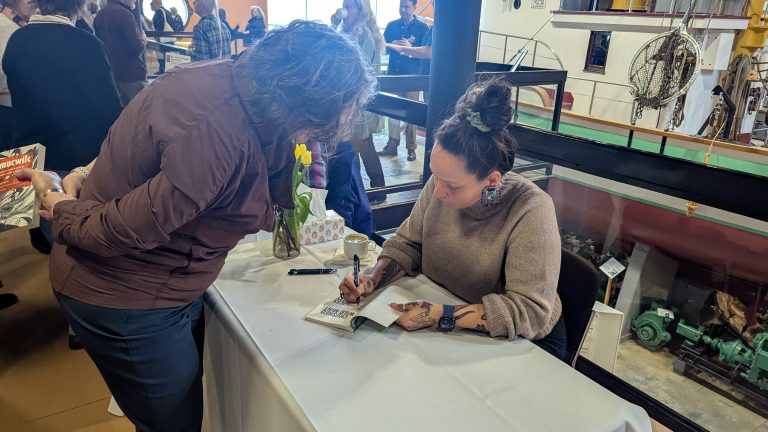 A woman sits at a table signing a book for another woman who is leaning over. People stand in the background near a display with machinery and a ship's interior. A vase with yellow flowers is on the table.