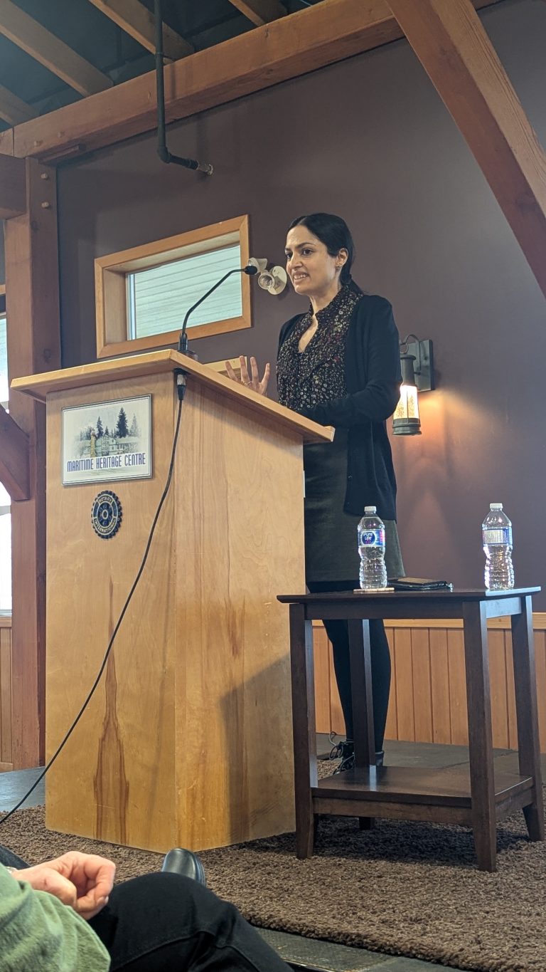 A woman stands at a wooden podium, speaking into a microphone at the Maritime Heritage Centre. She gestures with her hands. Two water bottles are on a small table beside her.