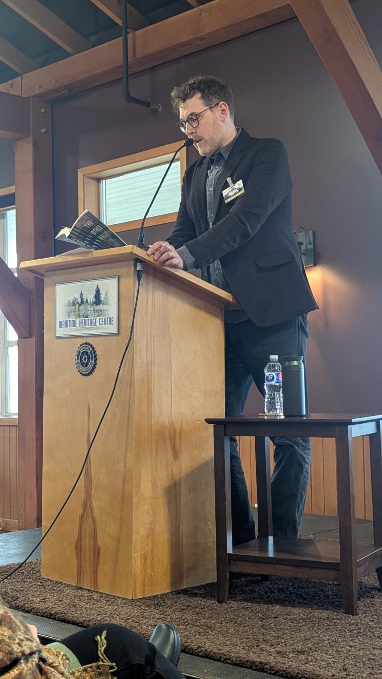 A man in a suit reads from a book at a wooden podium with a microphone in a rustic room. A water bottle and mug are on the podium, and people are seated in front of him.