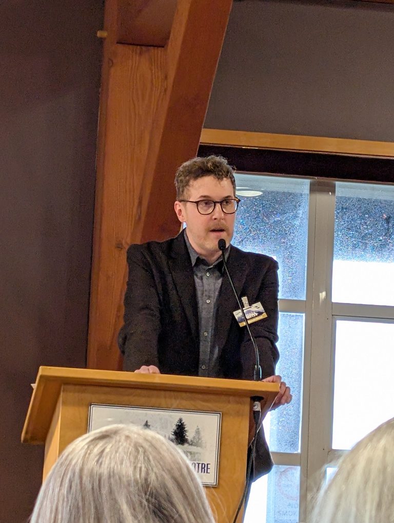 A man in a dark suit and glasses speaks at a wooden podium with a microphone, indoors near a window. A sign with trees is on the podium. The backs of people’s heads are visible in the foreground.
