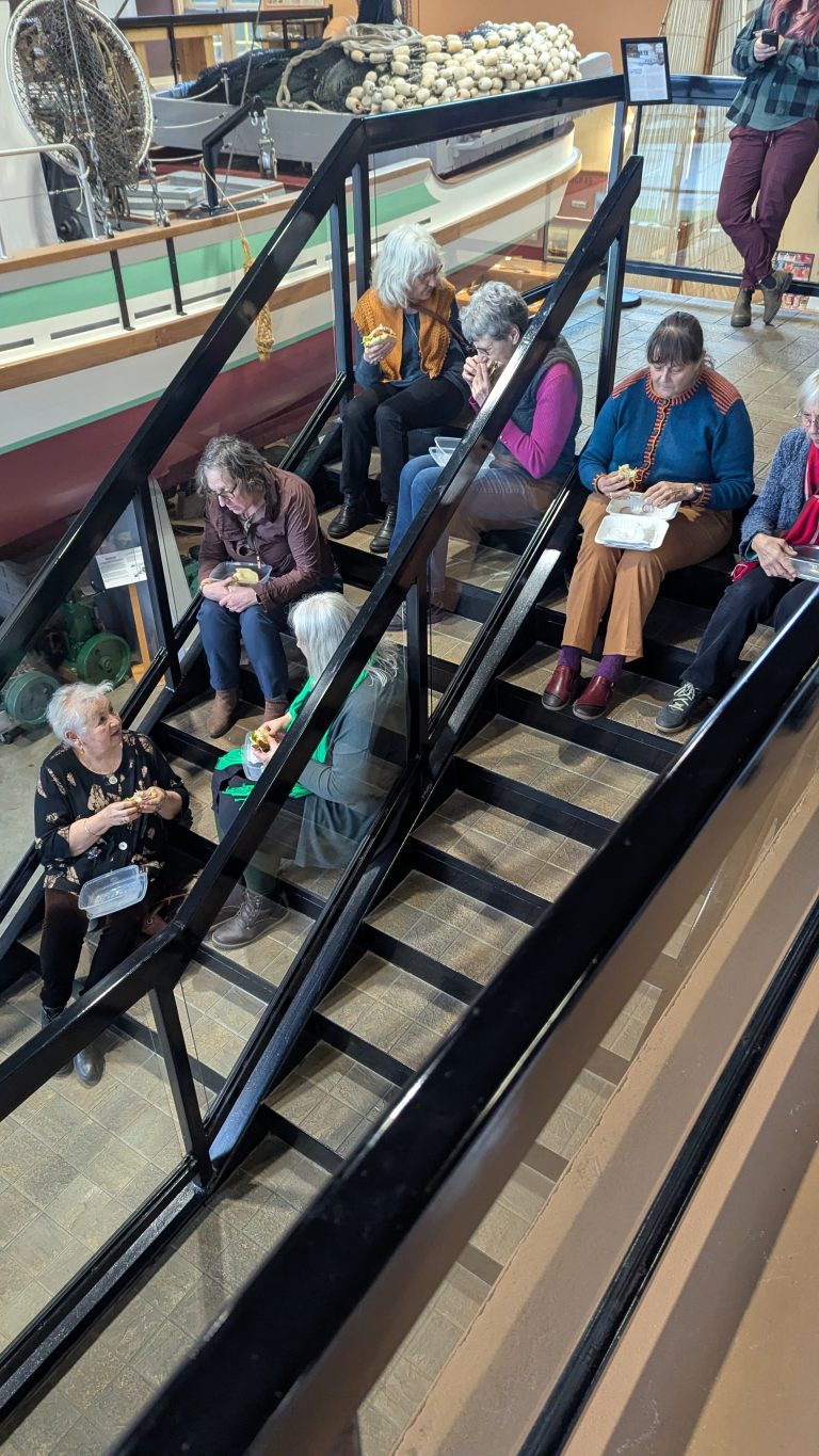 A group of older adults sit on a staircase indoors, eating and talking together. Some hold plates of food. There is a boat and other museum displays in the background.