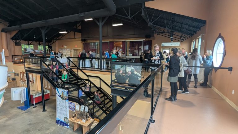 A group of people gather inside a spacious, modern building with high ceilings, black railings, and large windows. Some stand on an upper level, while others are on stairs and the lower floor, engaging in conversation.