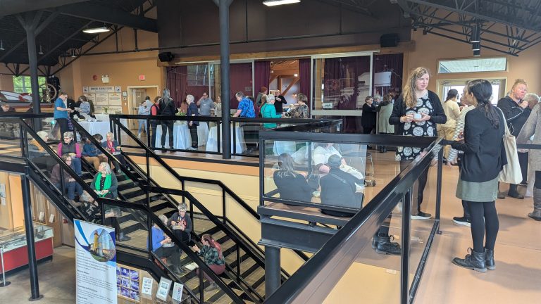 People gather and converse in a spacious, two-level indoor venue with large windows, staircases, and informational displays. Some stand in groups, while others walk or sit throughout the area.