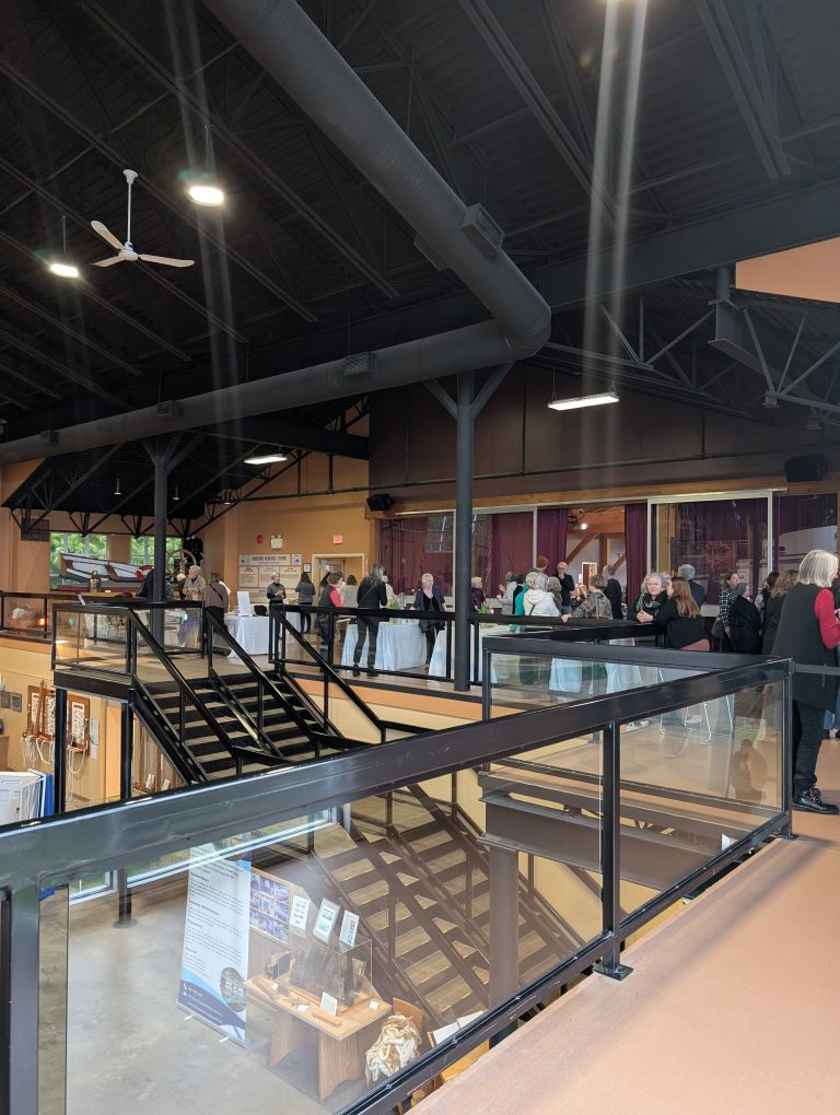 People gather in a spacious indoor venue with high ceilings, large windows, and exposed beams. Some stand near tables along a railing overlooking a lower level with stairs and display boards.