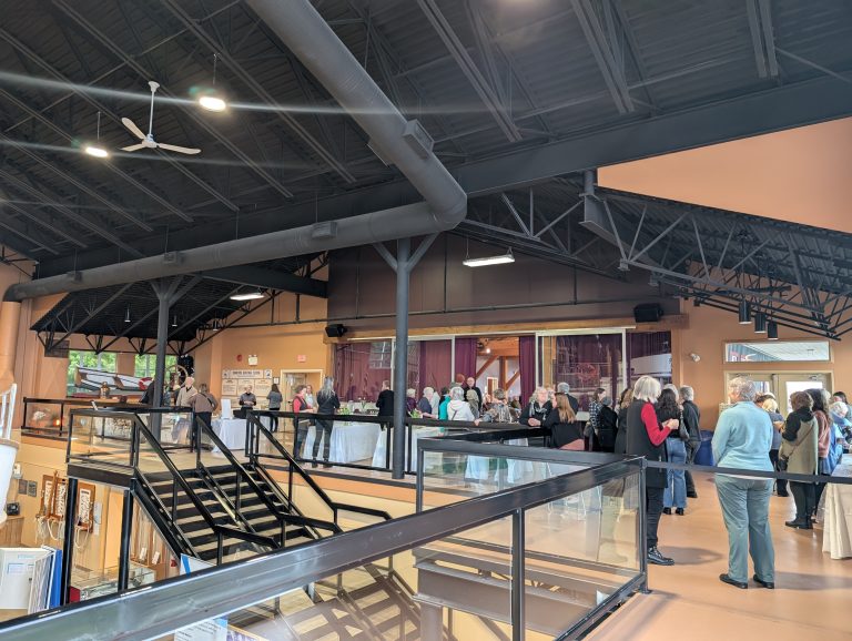 A group of people gathers inside a spacious building with high ceilings, near tables and displays. Large black beams, stairs, and glass railings are visible, with sunlight coming through the windows.